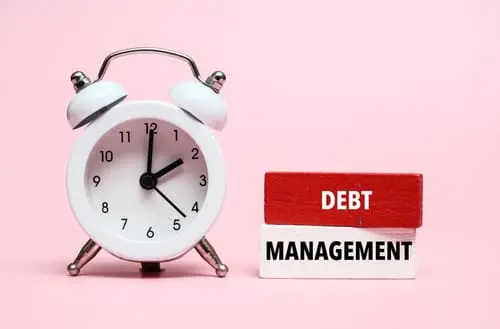 A coloured wooden block with text DEBT MANAGEMENT on it next to white alarm clock.