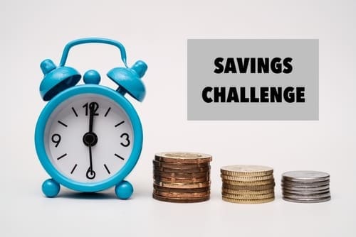 A blue alarm clock next to stacks of coins and a sign that reads SAVINGS CHALLENGE. 