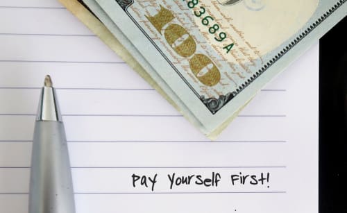 A pen-on-paper notebook with cash, dollars, and money is all on white-lined note paper with text written PAY YOURSELF FIRST.