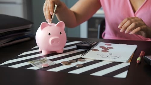 A young woman uses a calculator to count coins and banknotes while sitting at a kitchen table, placing money into a pink piggy bank.