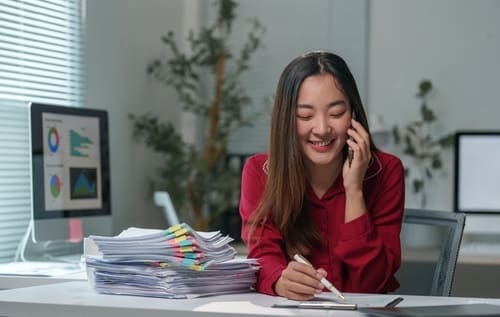 Young office worker smiling and talking on the phone while taking notes, with a large pile of paperwork and a computer with charts on her desk