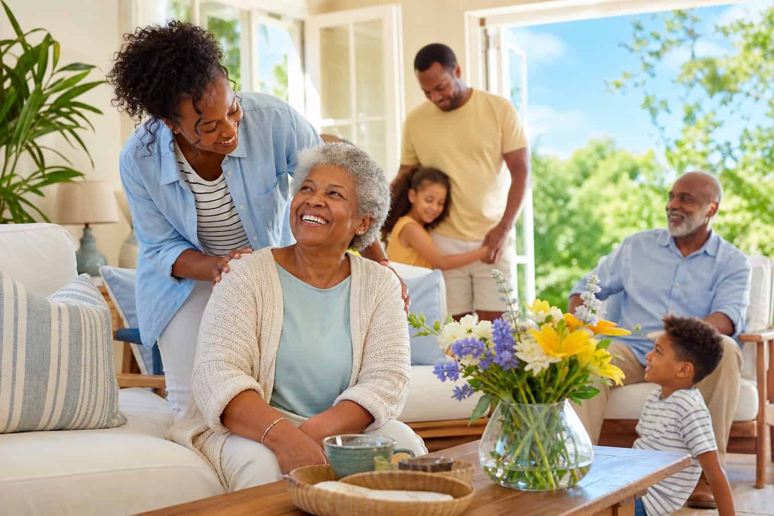 A multi-generational family in a sunlit living room, with a younger adult assisting an elderly person, surrounded by summer flowers and open windows showing green trees.